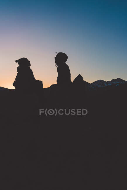 Silueta de hombre y mujer relajados sentados en retroiluminación en la cima de la montaña al atardecer - foto de stock