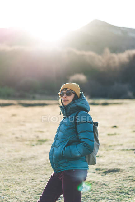 Вид сбоку на молодого путешественника в теплой активной одежде с рюкзаком и солнцезащитными очками, пересекающими пустые сухие травы — стоковое фото