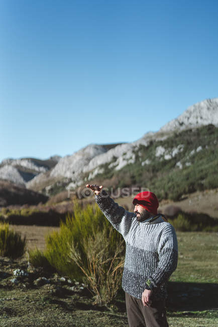 Vista laterale del viaggiatore maschio adulto barbuto in maglione grigio e berretto rosso a maglia in piedi con mano rialzata nella valle di montagna — Foto stock