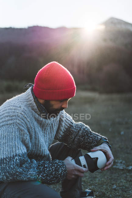 Вид збоку бородатого чоловіка-фотографа в червоному в'язаному в'язанні захоплює професійну камеру в рюкзаку в горах — стокове фото