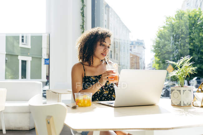 Mulher étnica com cabelo encaracolado bebendo suco fresco e usando laptop enquanto sentado contra a janela no moderno restaurante elegante — Fotografia de Stock