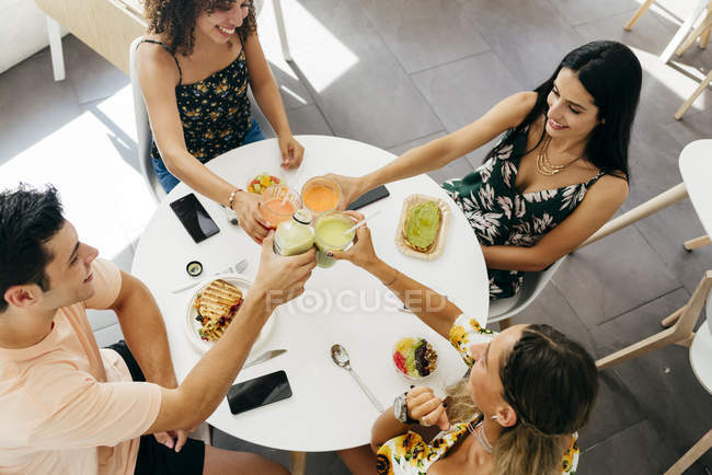 Young people clinking glasses of fresh fruit beverages and proposing toast while sitting at table with healthy snacks in cafe — Stock Photo