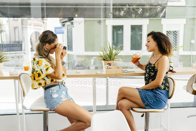 Side view of young lady taking photo of ethnic female friend with healthy drink in stylish restaurant — Stock Photo