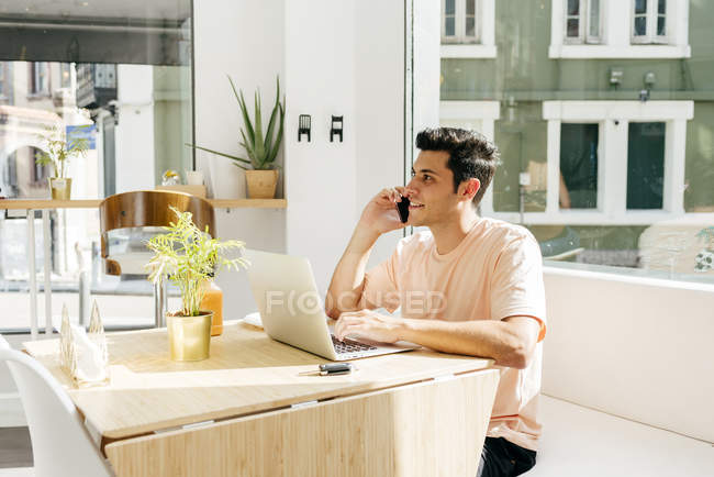 Hombre joven usando el ordenador portátil y hablando de teléfono en la cafetería - foto de stock