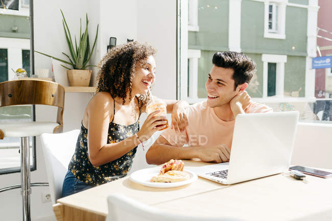 Mujer étnica alegre disfrutando de bebidas frescas y apoyándose en el hombro del novio con el ordenador portátil mientras está sentado en la mesa de la cafetería - foto de stock