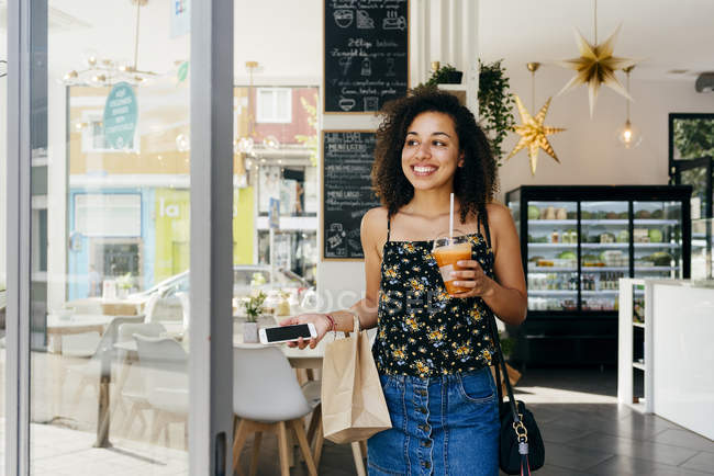Mujer étnica feliz con teléfono inteligente y jugo fresco sonriendo y mirando hacia otro lado mientras deja la cafetería moderna - foto de stock