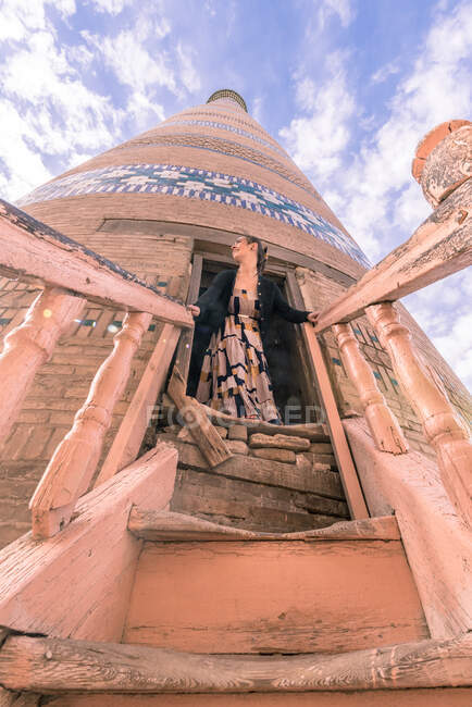 Снизу женщина-туристка в повседневной одежде стоит и смотрит в сторону в дверях древнего высокого коричневого кирпича Ислам Ходжа минарет над старой деревянной обветшалой лестницей в Хиве под голубым небом — стоковое фото
