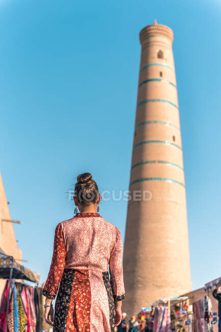 Back view of female in colorful dress walking along open air oriental market with traditional clothes and souvenirs near tall brown brick tower of Islam Hodja minaret in Khiva under cloudless blue sky — Stock Photo