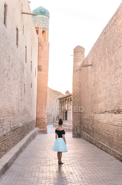 Vista trasera de la mujer en falda romántica de pie solo en la calle estrecha contra edificios antiguos marrones con paredes de adobe en Khiva Uzbekistán - foto de stock