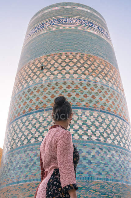Desde abajo vista trasera de la señora en ropa tradicional de pie solo y mirando la torre redonda incompleta del minarete Kalta Menor con colorido muro de ladrillo brillante en la calle en Khiva - foto de stock