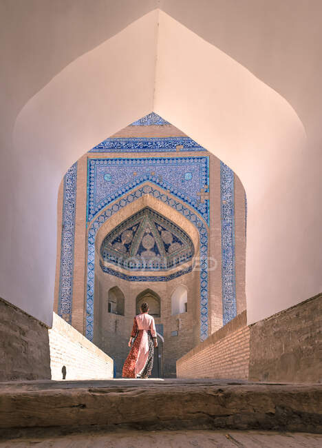 На задньому плані жінка в барвистому вбранні йде по вулиці до старого будинку з аркою і блакитною мозаїкою на стінах Хіви в погоду. — стокове фото