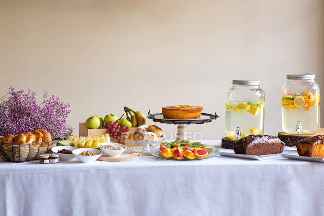 Vista lateral de la cena festiva con sabrosa comida de panadería de frutas y elegantes bancos de limonada en la mesa decorada con flores de lila. - foto de stock