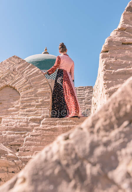 Снизу сзади вид спокойной женщины в традиционном длинном платье, стоящей на каменной лестнице и смотрящей на старый голубой купол мавзолея Пахлавана Махмуда в Хиве — стоковое фото