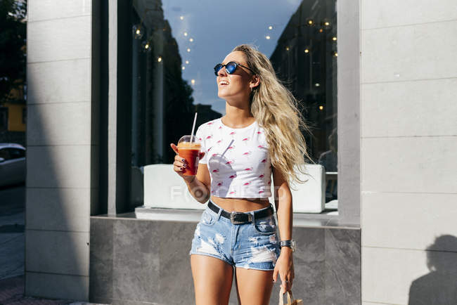 Весела жінка в модному вбранні зі свіжим напоєм, який дивиться далеко, стоячи на міській вулиці — стокове фото