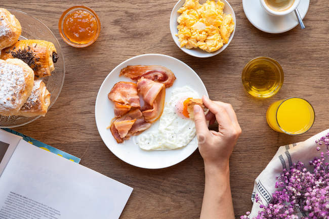 De cima da pessoa de cultura que molha pão em gema de ovos fritos com bacon para o café da manhã e livro de leitura na mesa servida — Fotografia de Stock
