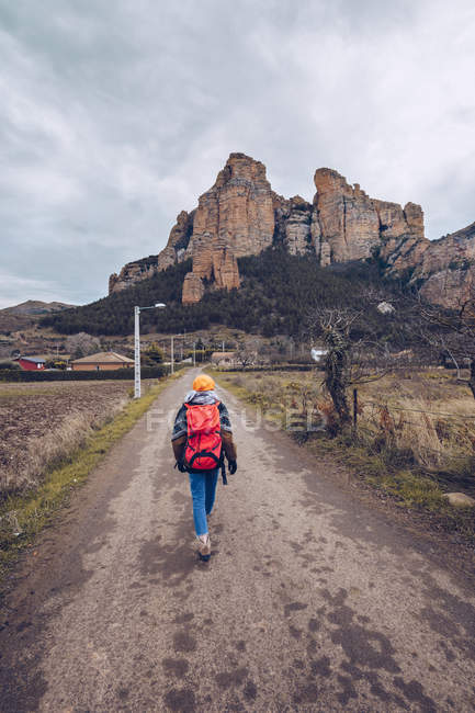Da dietro escursionista con zaino a piedi lungo un sentiero di cemento con alberi spogli e montagne lontane — Foto stock
