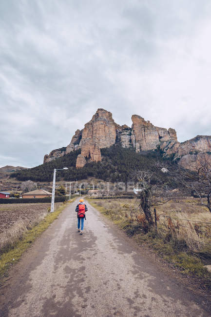 Von hinten Wanderer mit Rucksack auf Betonpfad am Feld mit kahlen Bäumen und weit entfernten Bergen — Stockfoto