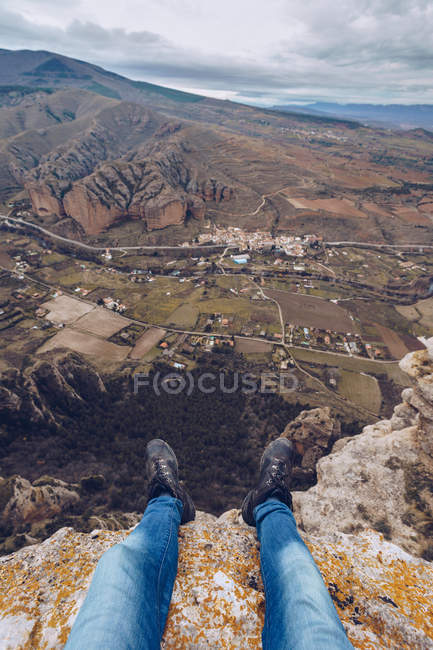 Pieds de personne en jeans assis sur le bord de la montagne avec vue imprenable sur la vallée pittoresque — Photo de stock
