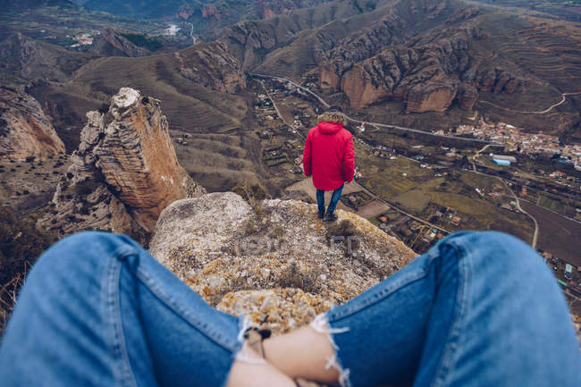 Recortado de mujer en jeans sentado con las piernas cruzadas, mientras que el hombre con chaqueta mirando y de pie en la cima de las montañas - foto de stock