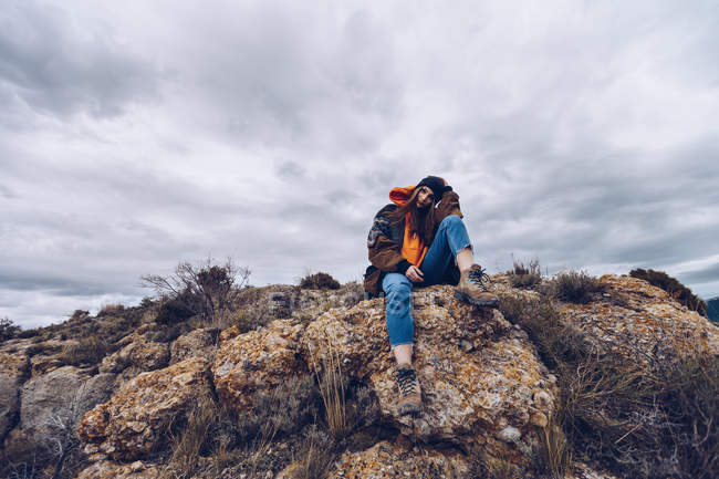 Dal basso donna positiva guardando in macchina fotografica e seduto su rocce sul bordo della scogliera con vista pittoresca — Foto stock