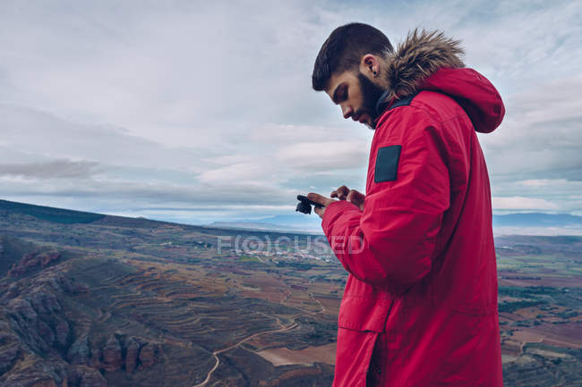 Side view of attentive man in jacket using smartphone while standing at cliff over breathtaking view — Stock Photo