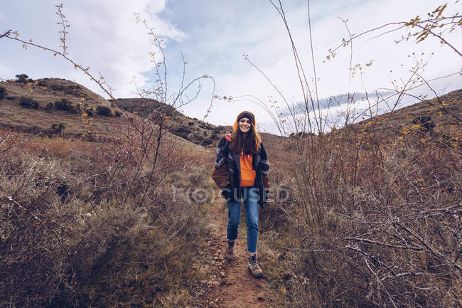 Jovem com mochila brilhante em roupas quentes andando ao longo da estrada com folhas secas por árvores nuas — Fotografia de Stock