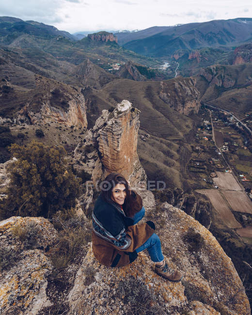 From above positive woman looking in camera and sitting on rocks at edge of cliff with picturesque view — Stock Photo