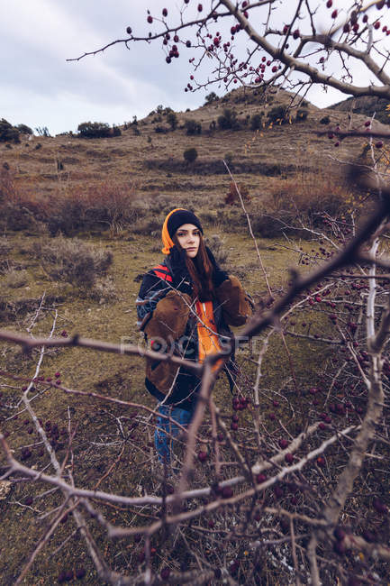 Junge Frau in warmen Kleidern steht mit getrockneten Blättern an kahlen Bäumen auf einem Feld und blickt in die Kamera — Stockfoto