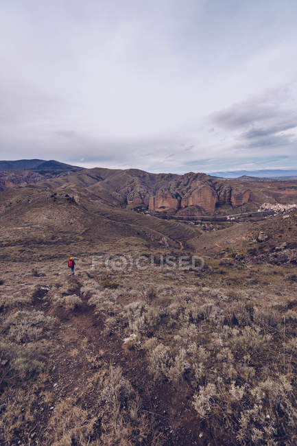 Отдаленный турист с ярким рюкзаком в теплой одежде, идущий вдоль поля с горами вдали — стоковое фото
