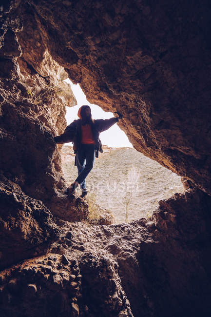 Mulher despreocupada em roupas quentes em pé à entrada do buraco rochoso à luz do dia — Fotografia de Stock