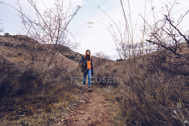 Молода жінка з яскравим рюкзаком в теплому одязі, що йде по дорозі з сушеним листям оголеними деревами — стокове фото