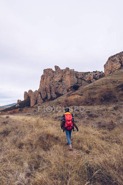 From behind hiker with bright backpack in warm clothes walking along concrete pathway at field with distant mountains — Stock Photo