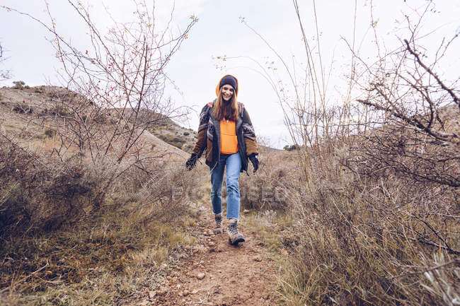 Mujer joven en ropa de abrigo caminando por el camino con hojas secas por árboles desnudos - foto de stock