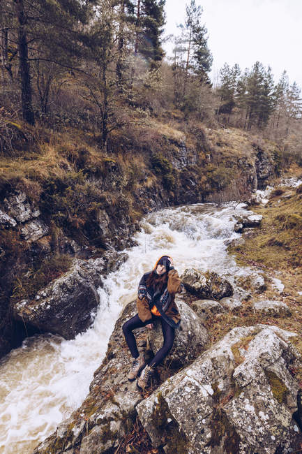 Женщина в теплой куртке сидит в яростном потоке в вечнозеленом лесу холодным осенним днем и смотрит в камеру — стоковое фото