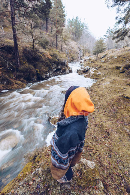 From above woman in warm jacket standing by furious torrent at evergreen forest on cold autumn daytime — Stock Photo