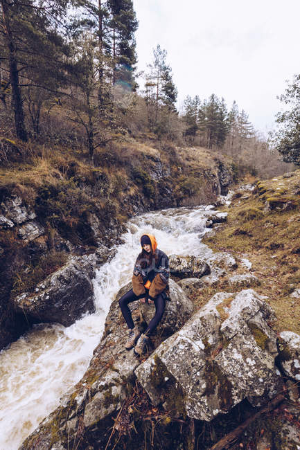 Donna in giacca calda seduta da torrente furioso a foresta sempreverde nella fredda giornata autunnale e guardando in macchina fotografica — Foto stock