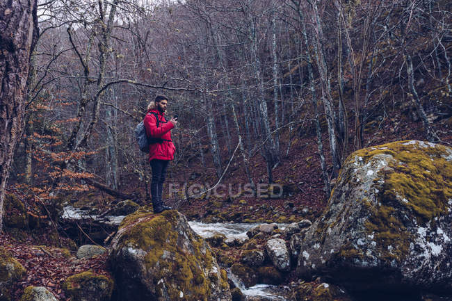 Вид збоку бородатого чоловіка з рюкзаком серфінгу смартфон, стоячи на скелях біля гірської річки — стокове фото