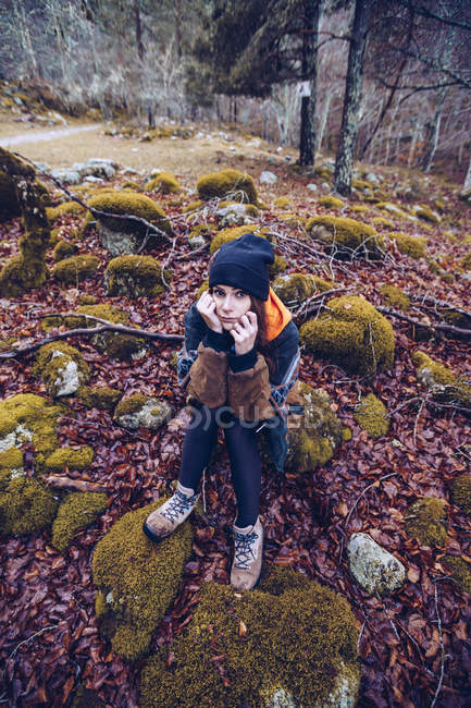 From above woman in hat leaning on hands while sitting in stones surrounded with dried foliage and moss — Stock Photo