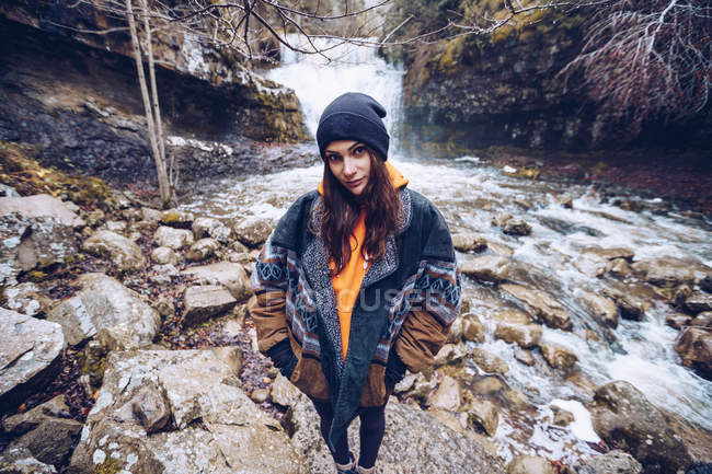 Женщина в теплой куртке, стоящая у бешеного потока в вечнозеленом лесу холодным осенним днем и смотрящая в камеру — стоковое фото