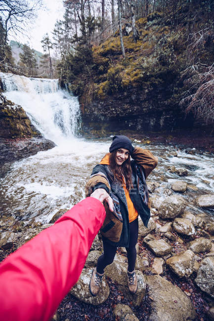 Mujer en ropa de abrigo caminando y cogida de la mano del hombre por estanque frío con cascada en bosque siempreverde - foto de stock