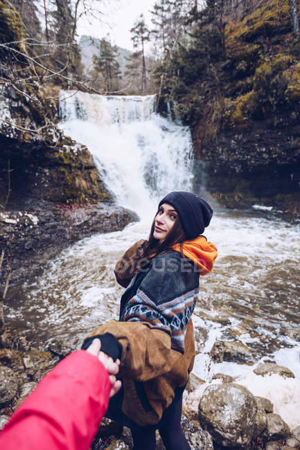 Frau in warmen Kleidern geht und hält Hand des Mannes am kalten Teich mit Wasserfall im immergrünen Wald — Stockfoto