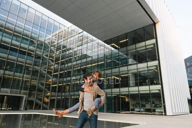 Happy man carrying woman piggyback while standing and laughing on street against contemporary business center — Stock Photo