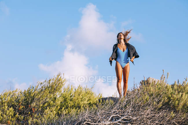 Contended tanned lady in trendy striped swimsuit walking on countryside hills against blue sky — Stock Photo