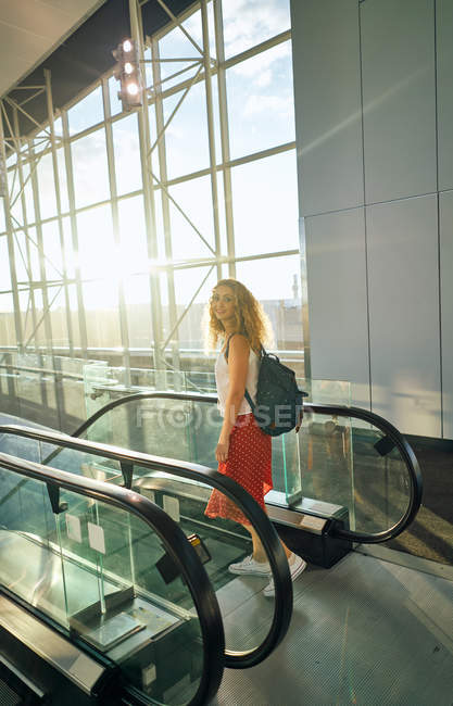 Vista lateral da encantadora mulher encaracolada em saia vermelha com mochila andando para baixo na escada rolante no aeroporto no Texas — Fotografia de Stock