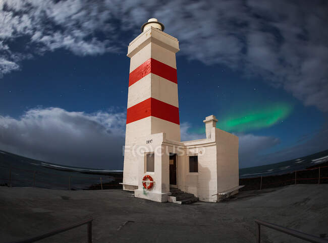 Weißes Leuchtfeuer mit gestreiftem Turm an der Küste vor blauem Nachthimmel mit Wolken und Nordlichtern in Island — Stockfoto