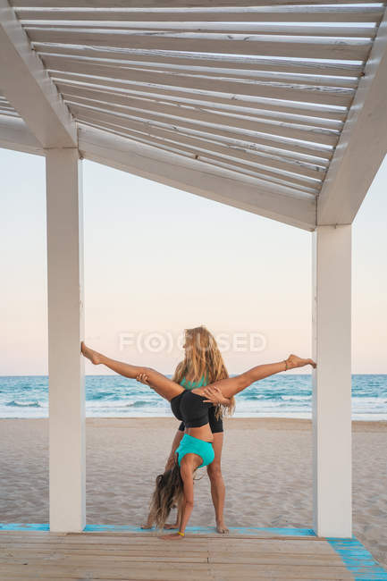 Vista lateral de mulheres esportivas ativas preparando-se para acroyoga posição em suporte de madeira na praia arenosa — Fotografia de Stock