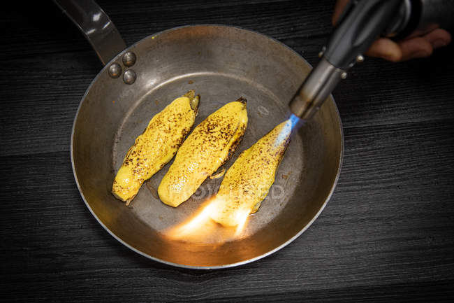 Рука людини, яка направляє пальника на сковороду з соковитими кінцями, готується на сковороді на сірому фоні — стокове фото