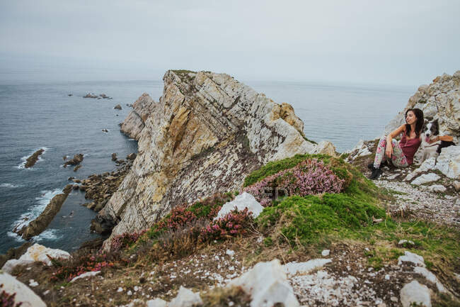 Mujer adulta con ropa casual mirando hacia otro lado y disfrutando de las vistas mientras está sentada y abrazando al perro Border Collie en la costa rocosa contra las olas grises bajo el cielo nublado en Asturias en verano - foto de stock
