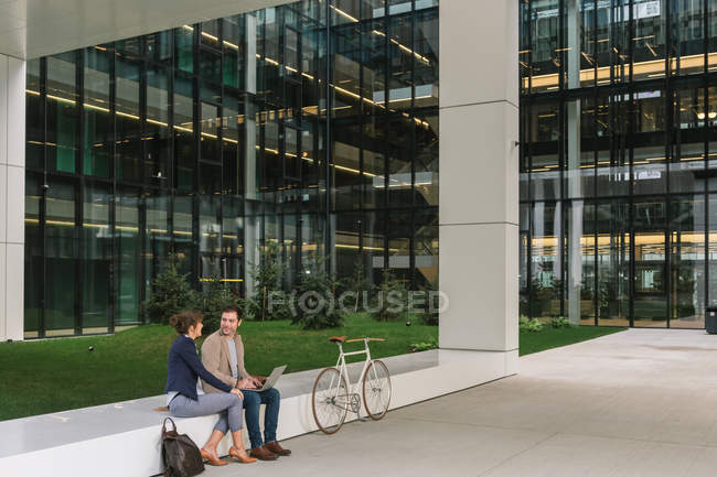 Delighted business people smiling and browsing laptop together while sitting outside modern building near bicycle on city street — Stock Photo