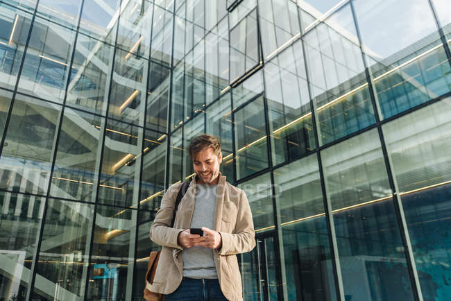Низький кут щасливого бізнесмена, який використовує смартфон, стоячи за межами бізнес-центру на міській вулиці — стокове фото